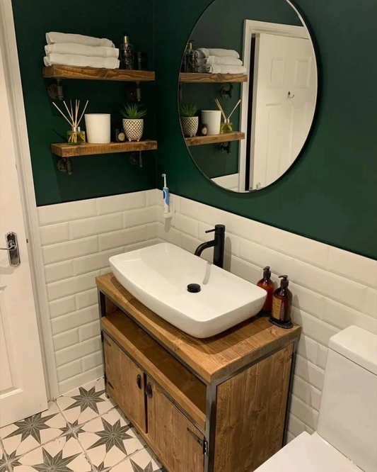 A rustic bathroom vanity unit with a wooden top and steel frame, positioned against a dark green wall with a white tiled floor, featuring a white sink and a round mirror above it.