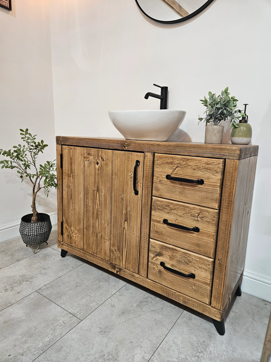 A rustic pine finish vanity unit with three drawers and a cupboard, featuring a vessel sink on top and a black tap. The unit is positioned on a tiled floor against a white wall, with a decorative plant and mirror in the background.