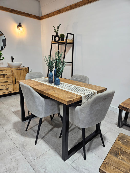 A rustic dining table with square metal legs and a wooden tabletop, surrounded by gray upholstered chairs in a well-lit room with decor.